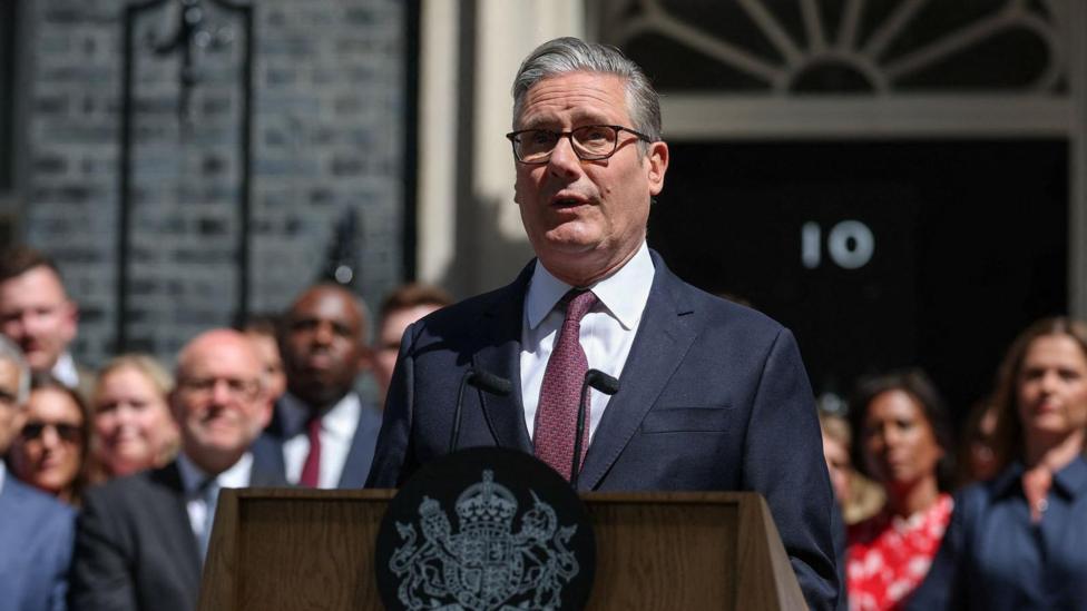Keir Starmer wears a suit, glasses and has short grey hair. He stands at a lectern outside 10 Downing Street with a crowd of people behind him