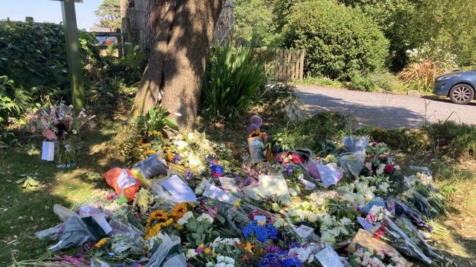 Dozens of bouquets of flowers laid on the grass in front of a tree near a hotel. Among the floral tributes at the back is a black and white photo of Widdecombe.