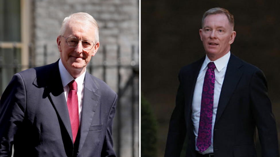 Two men in suits. On the left Hilary Benn has grey hair, dark rimmed glasses, a dark suit with a red tie and white shirt. He is standing in front of a building. On the right is Sir Chris Bryant, he is also wearing a dark suit, with a white shirt and purple patterned tie.
