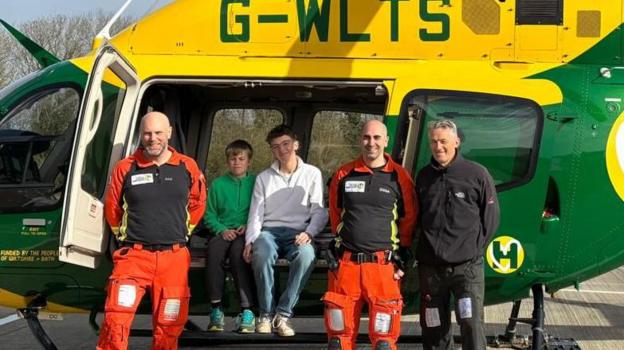 People in front of a colourful air ambulance helicopter, which is painted green and yellow. The helicopters side doors are open with two teenage boys sitting smiling at the camera. Standing beside them are three men - two in uniforms of orange and black and other dressed in black fleece and trousers.