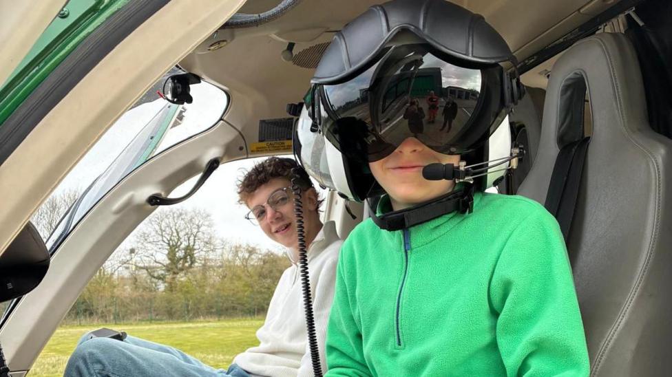 Two teenage boys smile from the cockpit of a helicopter on the ground. The one in the foreground wears a large pilots helmet with the visor down.