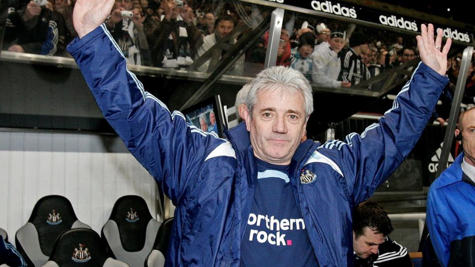 An old photo of Kevin Keegan when he was Newcastle manager in 2008. He has grey, short hair and is wearing a blue jacket with the Newcastle United logo and a blue top underneath with the Northern Rock sponsor peaking through. He is standing in front of the dugout at St James' Park and fans on the stands behind him. He has his hands in the air.