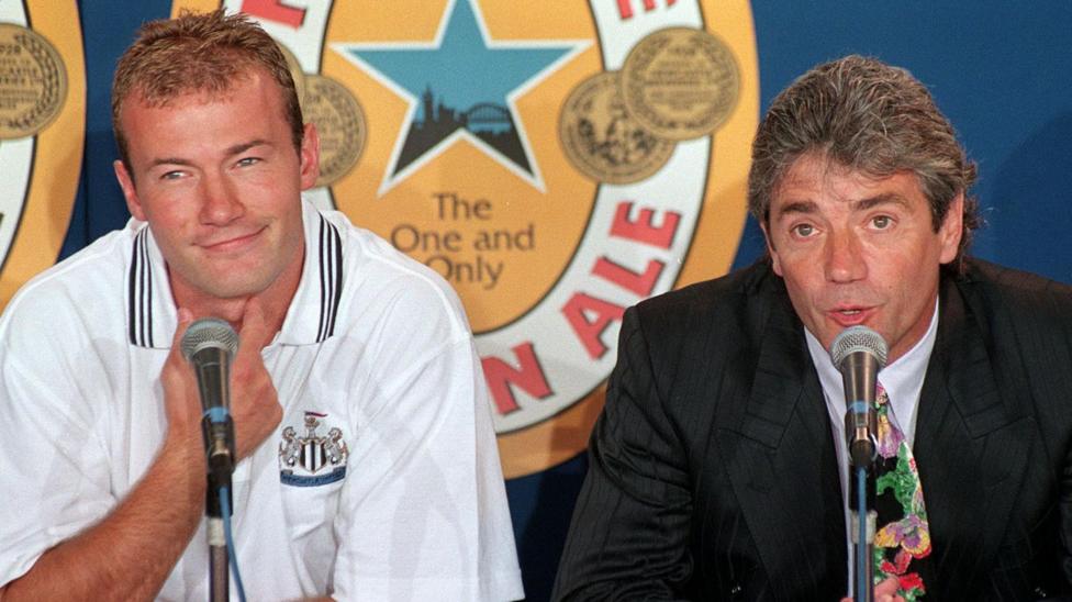 Alan Shearer sitting to the left of Kevin Keegan during a press conference. There are microphones in front of them both and Keegan is talking while Shearer looks off to the side and smiles. Shearer is wearing a white Newcastle United polo shirt while Keegan wears a black blazer and patterned tie. Shearer has dark blonde hair and Keegan has grey hair which is swept back. They are sitting in front of a large Newcastle Brown Ale advertisement.