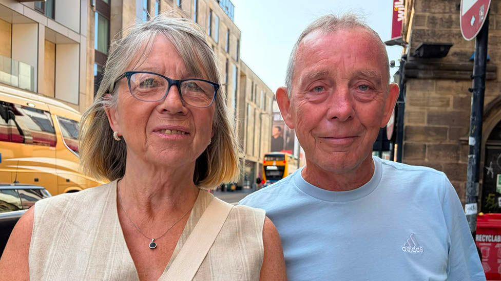 Brenda and Eddie smiling into the camera. Brenda is on the left and has short blonde hair and is wearing dark framed glasses with a beige striped top on. Martin has grey short hair and is wearing a blue t-shirt. They are standing on a busy street with buses going past.