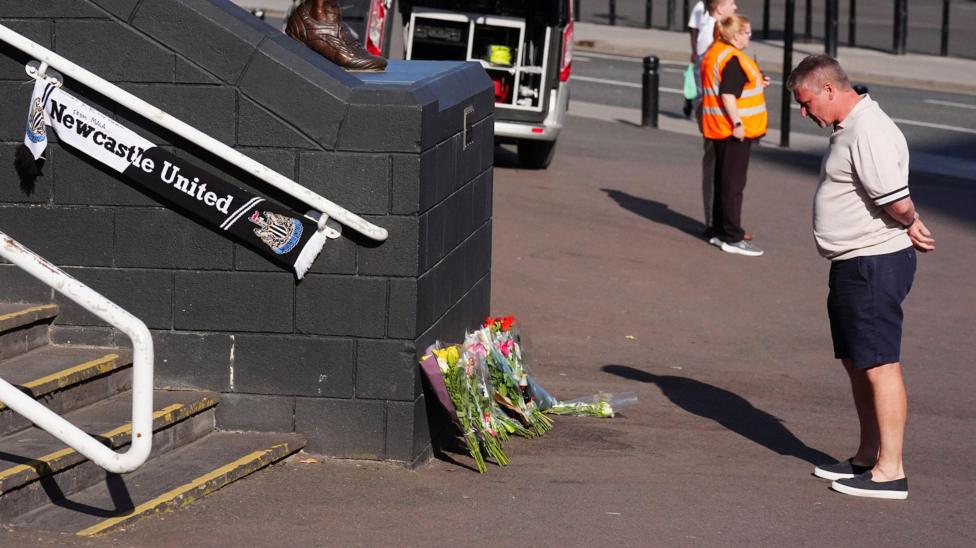 Lee Dixon standing in front of a row of flowers at the steps of St James' Park. He has grey, short hair and is wearing a beige polo shirt and blue shorts. He has his hands behind his back and his head bowed. There is a Newcastle United scarf attached to the railing to the left.