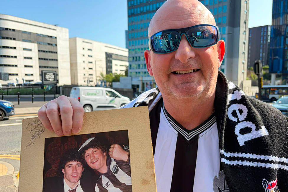 Malcolm smiling into the camera while holding up an old framed photo of himself and Kevin Keegan where Malcolm is wearing a curly dark wig to match Keegan's hair. Malcolm is bald and wearing blue sporty sunglasses and an old Newcastle shirt and scarf. He is standing in front of St James' Park and the Strawberry Place road is behind him.