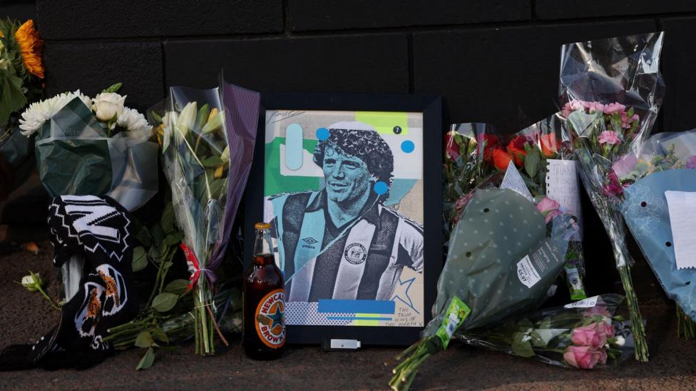 Bouquets of flowers leaning against a dark wall with a colourful print of Kevin Keegan when he was younger in a Newcastle shirt. There is a bottle of Newcastle Brown Ale next to it.
