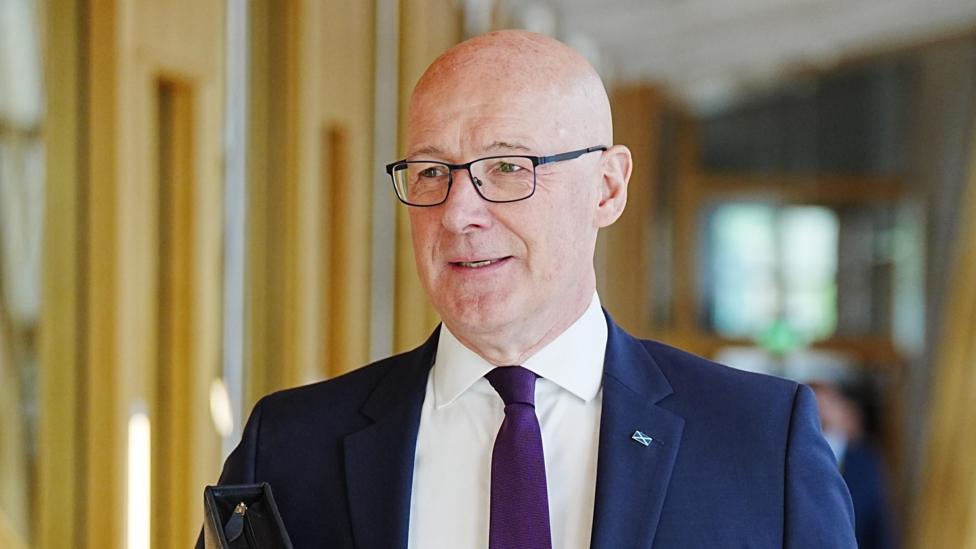 John Swinney wearing a blue blazer, purple tie, white shirt and glasses. He is walking down a corridor at Holyrood.
