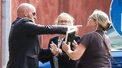 A woman holding a framed photo in front of a man. He is wearing a suit and is pointing behind her.