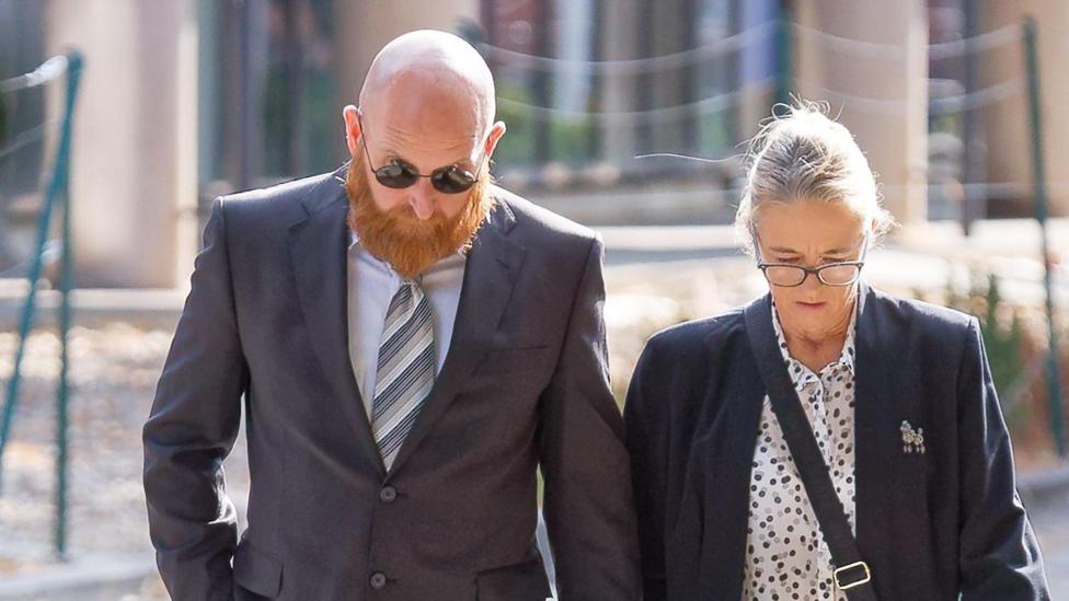 Neil Lewis wearing a suit and walking along with a blonde woman. There heads are both down.