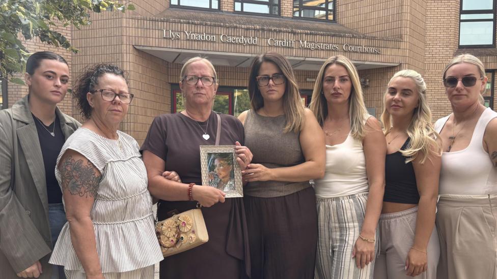 Seven woman stand together in a group. One is holding a framed photo of Darcy Ross. They all look at the camera.