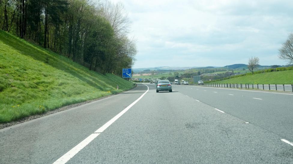 A stretch of motorway with a few cars on it and a blue sign in the distance. There are trees up a grass bank next to it and hills on the horizon.