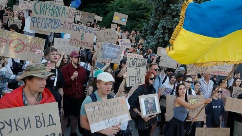 Dozens of people, mostly young, holding cardboard signs in a protest, as one woman waves a large Ukrainian flag