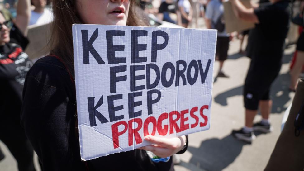A young woman holds a sign which says 'KEEP FEDOROV, KEEP PROGRESS'