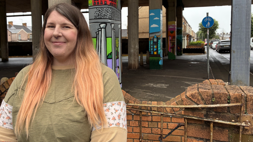 A woman with long blonde hair wearing a green top smiling in front of a wall and pillars that hold up the M4 with a road going under the motorway road deck
