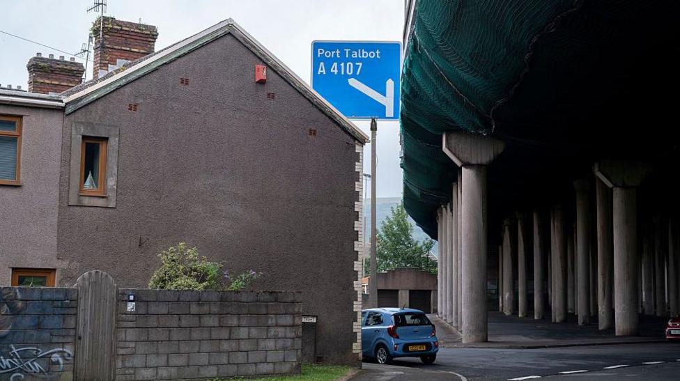 The images shows the side of a grey end-of-terrace house behind a breeze block wall with a blue car parked outside and next to the pillars that hold up the flyover of the M4 motorway at Port Talbot