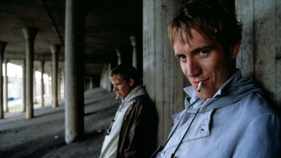 Two men with blond hair both smoking a cigarette standing underneath a motorway flyover with pillars in the background holding up the road deck