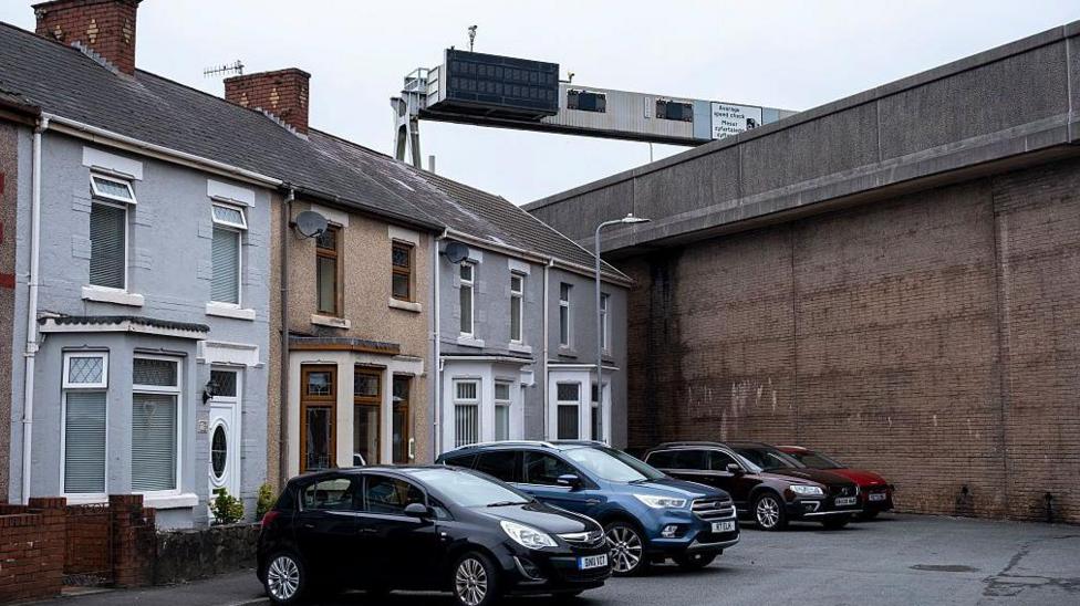 Cars parked outside houses in a cul-de-sac street in front of a large concrete wall which holds up the M4 motorway at Port Talbot