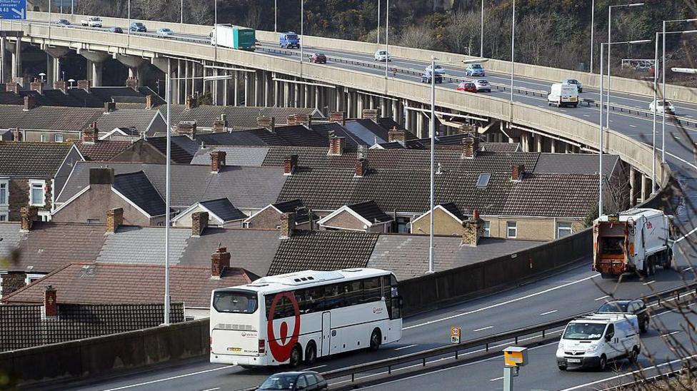 Cars, a bus, a dustbin lorry and vans on the M4 motorway flyover at Port Talbot as it passes by terraced houses