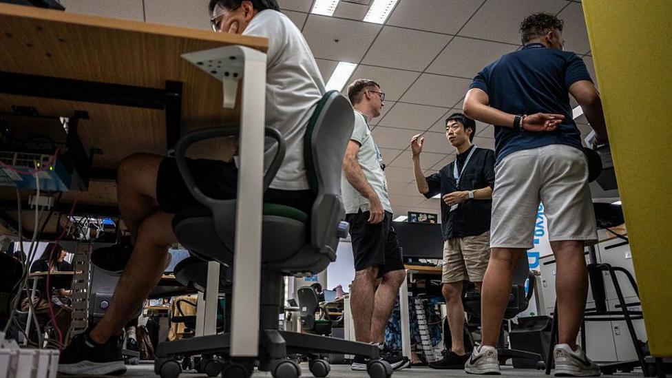 Four men wearing shorts in the office in Tokyo, Japan. Three of them standing up, one of them seated and with his elbows resting on his table.
