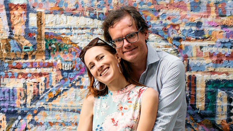 Photo of a couple with the man stood behind, hugging his partner. She wears a sleeveless floral top and sunglasses on her head, large gold hoop earrings, dark brown hair and he has dark brown hair, wears a blue and white striped top and spectacles. They both smile at the camera and are stood in front of a brightly coloured graffitied brick wall.