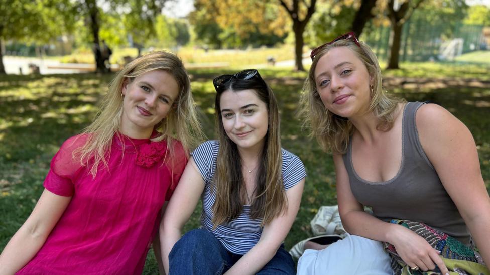 Group of three women sat in park, all smiling at the camera. The woman on left wears a fuchsia pink dress, the woman in the middle, a stripy t-shirt and jeans and then the woman on the right, a sage green vest top. Behind them is a park with leafy trees.