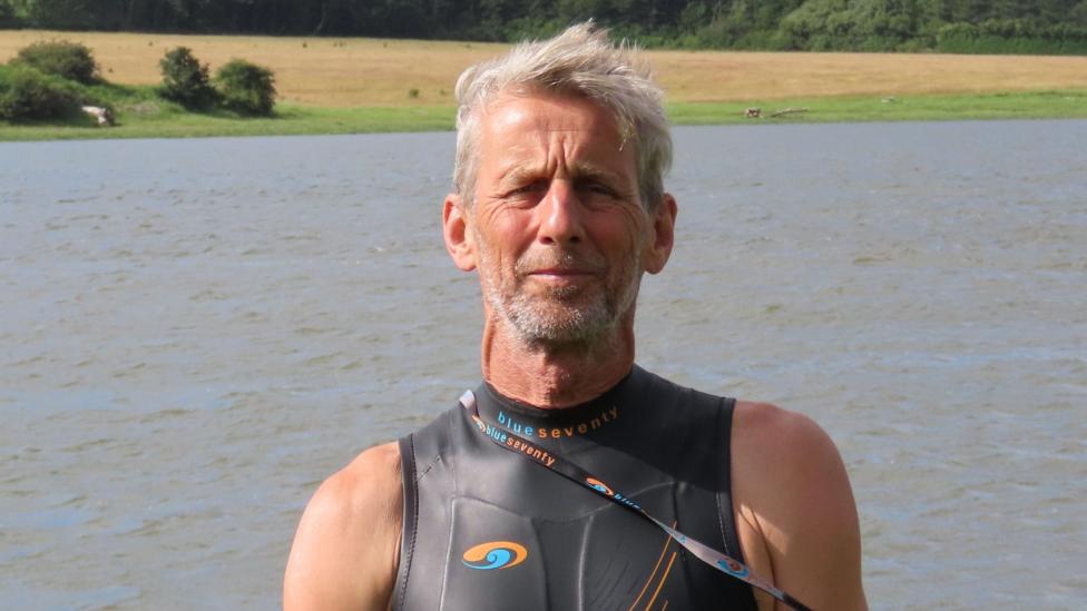 Ric Cooper of The Cleddau Project ready to go for a swim in the Western Cleddau river. He's standing in front of the river wearing a black, sleeveless wetsuit. He has grey short hair and grey stubble. It is a head and shoulders shot of him.
