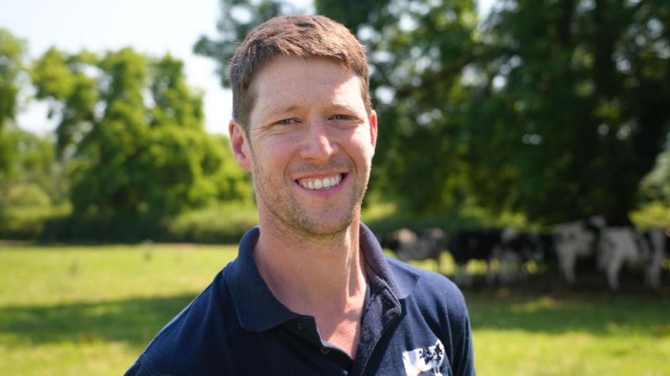 Dairy farmer Tom Jones stands in a field. He has short brown hair and is wearing a dark blue polo shirt with a black and white Holstein Friesian dairy cow logo. Behind him several cows can be seen seeking shade from the sun under a tree.