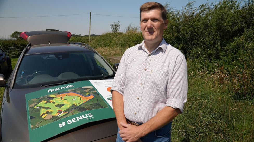 Lee Truelove of First Milk stands in front of a car in a field. On the bonnet is a large cardboard poster showing an example of one of the risk maps the company has produced for each of its farmers. It is a satellite image with fields colour-coded according to risk. Mr Truelove has short brown hair and is wearing a white shirt tucked into light blue jeans