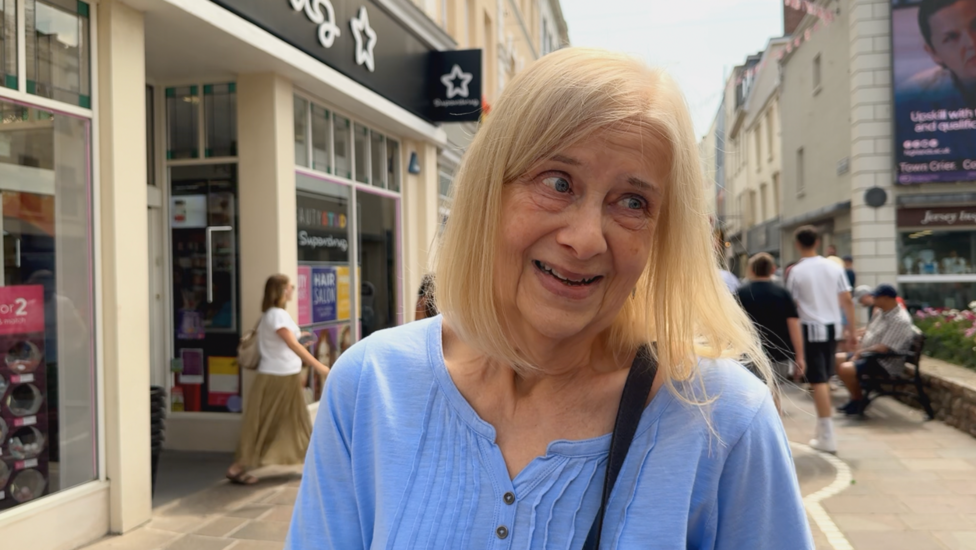 Interviewee Carole - a woman with shoulder-length light-coloured hair, wearing a light blue top and a cross-body bag. She is standing on a pedestrian Jersey's high street. Storefronts and signs line the street behind her, while several people walk or sit in the background.