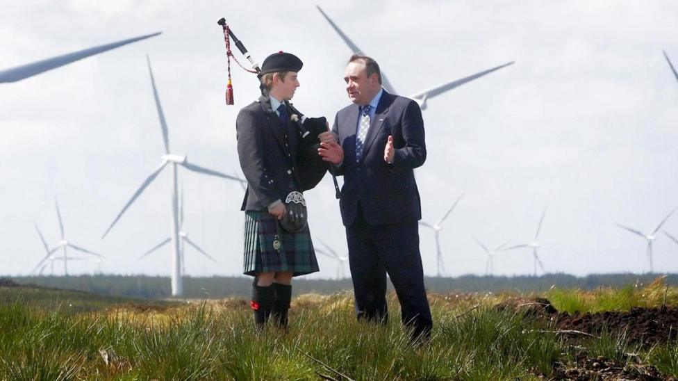 Former first minister of Scotland, Alex Salmond, is talking to a bagpiper. They are standing on grass in front of a large wind farm.