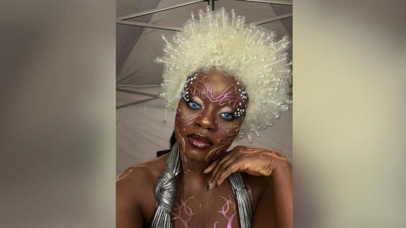 A person in bold avant‑garde makeup with bright blue eyes, a large white beaded wig, and metallic costume accessories, standing in a tent-like backstage setting