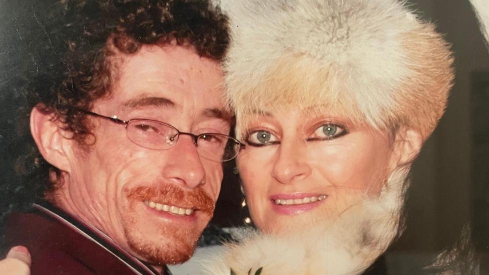 Man and woman hugging while smiling at the camera. Male has dark curly hair and a ginger beard wearing glasses. Female has white fluffy hat and scarf on.