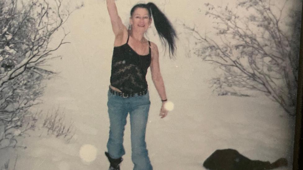Woman standing in the snow with one hand in the air. She is wearing a strap top and jeans. Her dark hair is in a high ponytail.