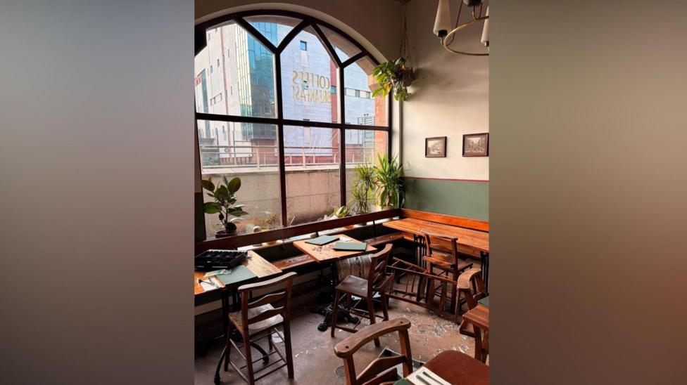 The inside of a coffee shop. The window is broken, and there is broken glass all over the floor of the coffee shop. There is a chair which appears to have been overturned.