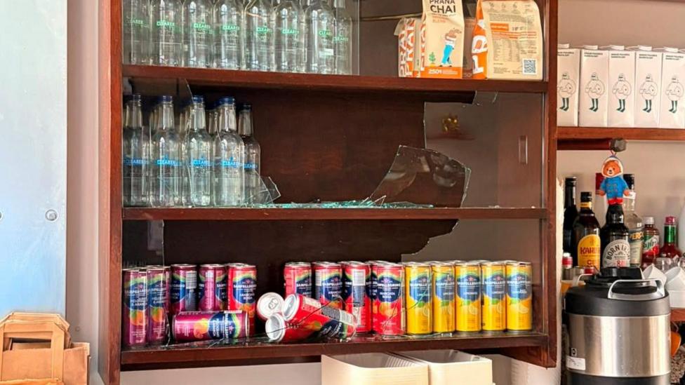 A wooden cabinet with soft drinks on several shelves. The glass window at the front of the cabinet is broken, with broken glass on some of the shelves.