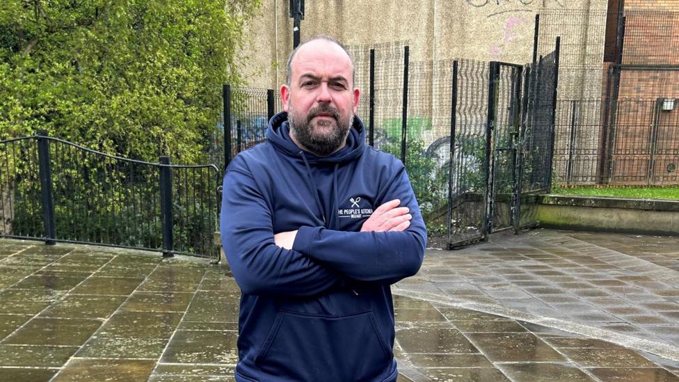 Paul McCusker is bald, he is wearing a navy hoody and dark blue tracksuit bottoms. He has his arms folded and is standing in a public square in Belfast.