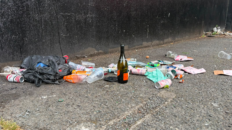 A collection of litter sits on a black concrete pavement. The litter includes cans, bottles, plastic cups and tissue paper.