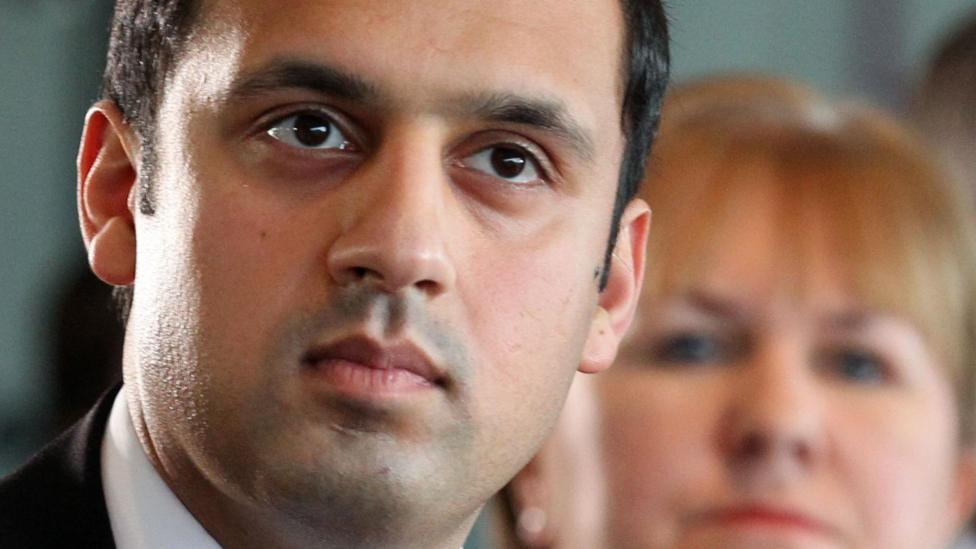 Anas Sarwar with a stern expression looking into the distance. He has black hair and looks to be wearing a suit. A woman with blonde hair is blurred in the background.