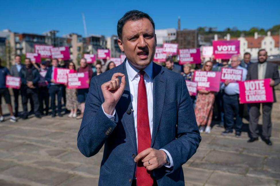 Anas Sarwar, mid speech with his fingers gesturing. He is standing in front of a crowd of people wearing a navy suit and red tie.