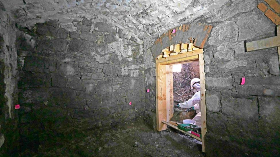 A person in white excavation/forensic attire kneeling beside a doorway. There are a number of pink stickers on the walls of a small building, which looks old and dilapidated.