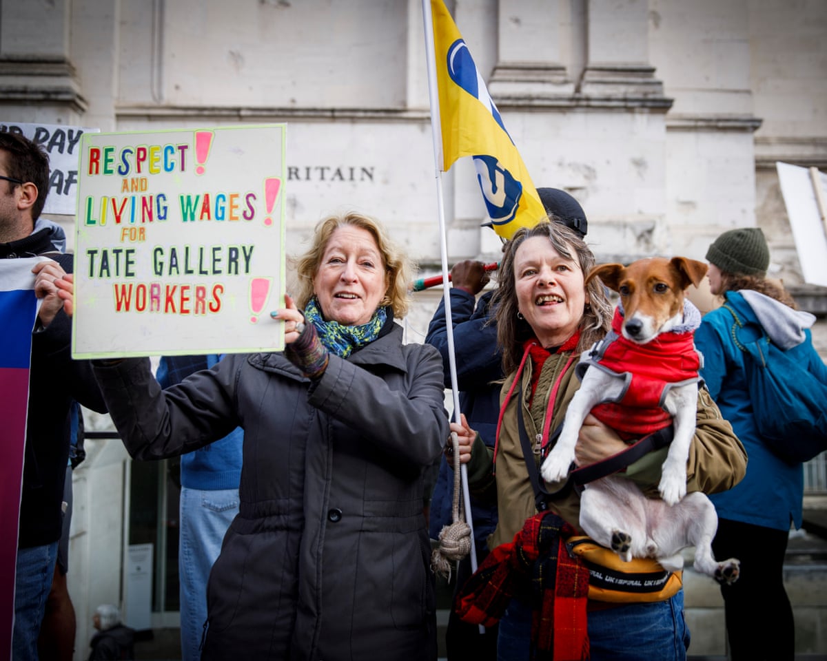 Two women strike, one holding a placard saying ‘Respect! Living Wages!’, the other holding a dog and flag.