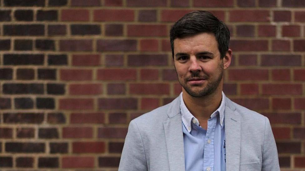 Seb Pelisse, who has dark, short hair and facial hair, stands in front of a brick wall smiling. He is wearing a blue shirt and light grey jacket.