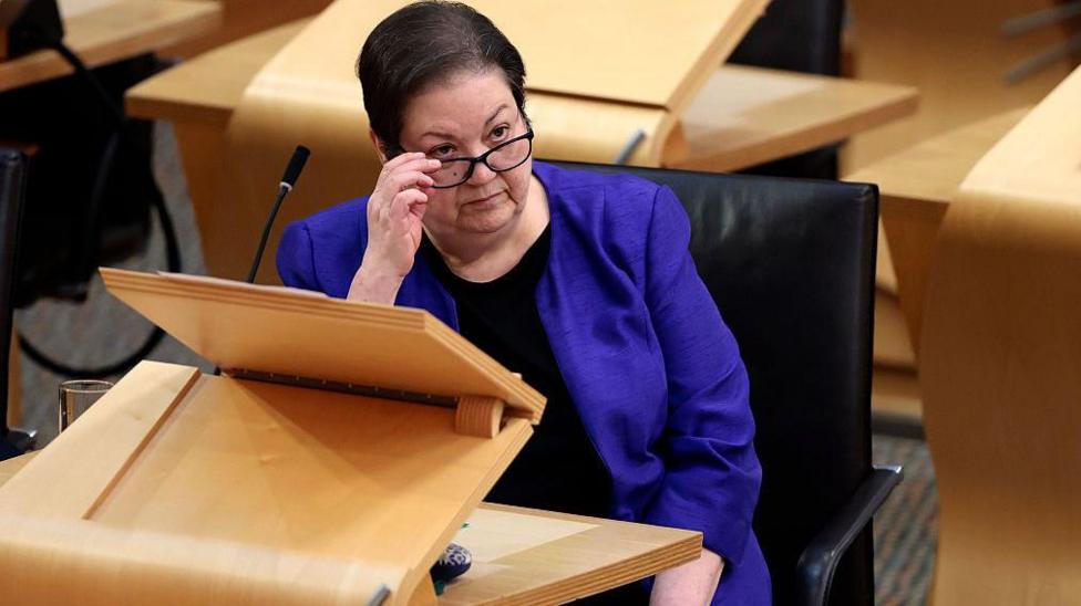 Jackie Baillie seated at a wooden desk in a parliamentary-style chamber, adjusting a pair of glasses. A microphone, black chair and surrounding wooden desks are visible.
