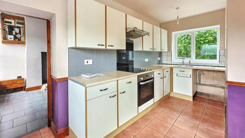 A kitchen with white kitchen units and purple walls. There is also brown/ orange tiles on the floor and blue and white tiles on the walls. 