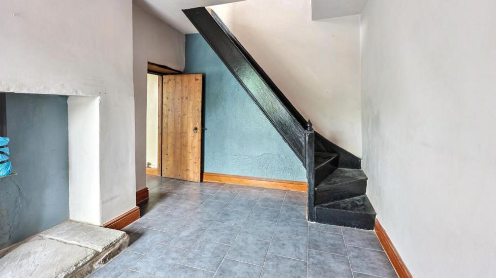 A grey tiled floor with a disused fireplace . There's black stairs in the corner. 