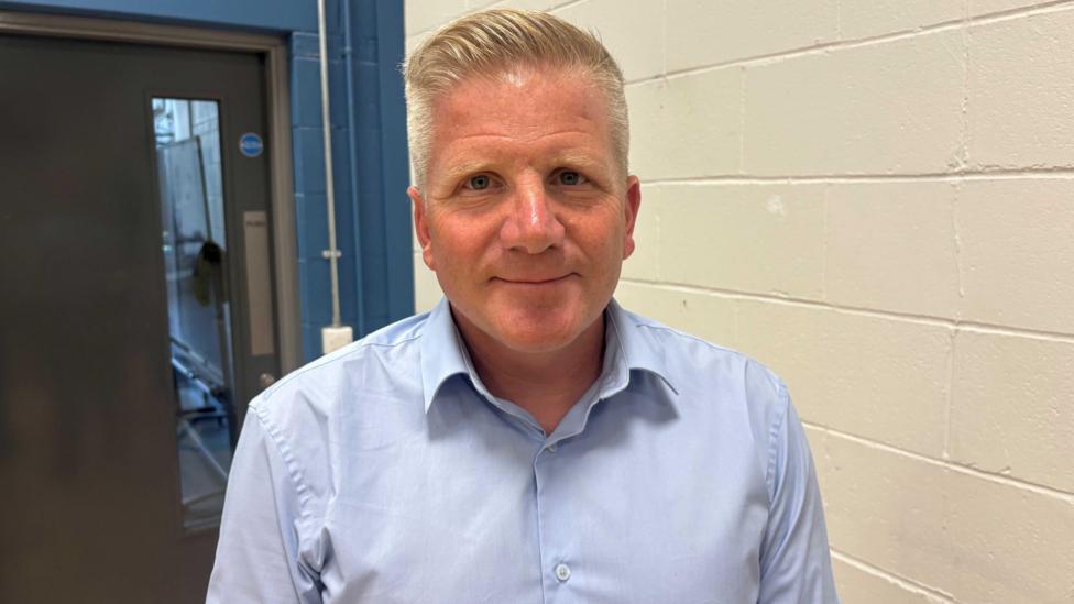 Chris Quinn, a man with short blond hair, smiles at the camera. He is wearing a light-blue shirt and is standing next to a white wall. Behind him is a door. 