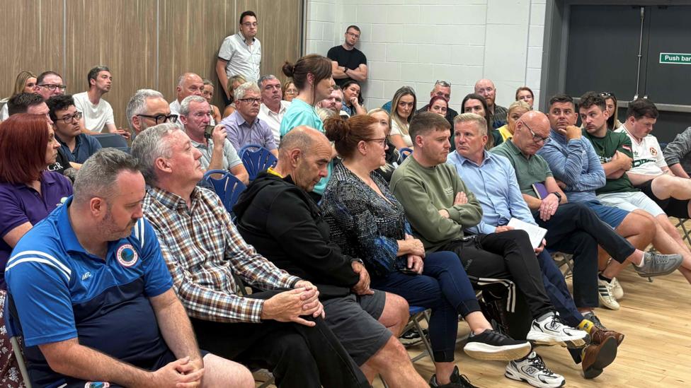 A large group of people sat on chairs. In the middle of the group, a woman in a cyan top is stood, seemingly talking. They are in a hall with a wooden floor in a community centre.