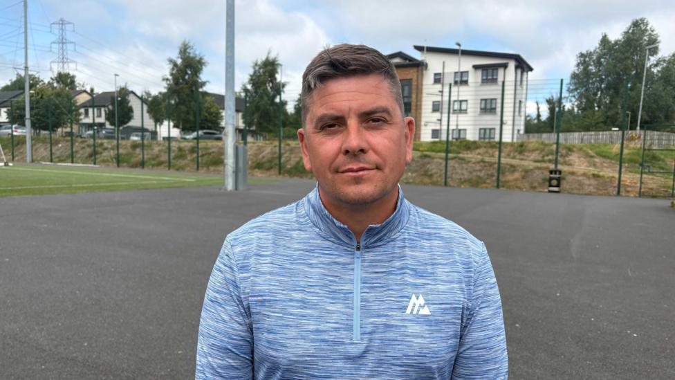 Danny Baker, with short dark hair, looks at the camera. He is wearing a light blue top. He is standing outside. Behind him is a large green fence, and behind that are some buildings. In front of the fence is a patch of grass with white sport-style markings on it. 