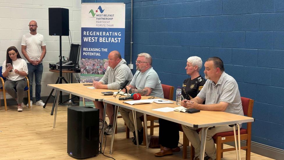 A tale with four people sat at it. On the table, there are a number of microphones, including a read microphone. The wall behind the table is dark-blue. On the left of the tale is a large sign which reads: 'REGENERATING WEST BELFAST - RELEASING THE POTENTIAL'. There is a man stood next t the sign, and a woman sat next to him. 